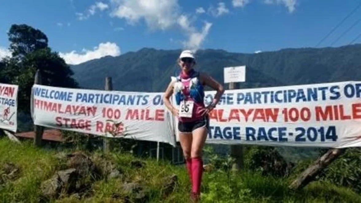 Una mujer está de pie junto a una pancarta que dice: "BIENVENIDOS PARTICIPANTES DE LA CARRERA POR ETAPAS DE 100 MILLAS DEL HIMALAYA - 2014".